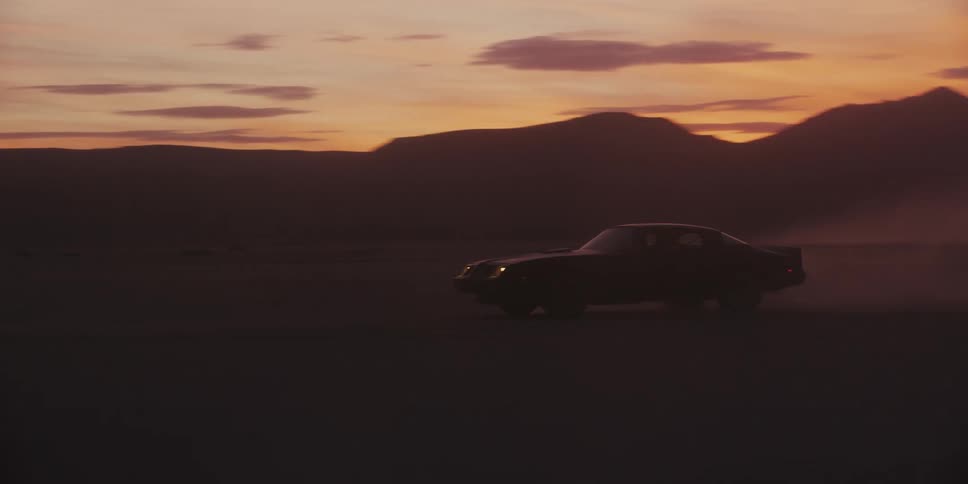 Black muscle car with headlights on driving on the desert near the mountains at sunset