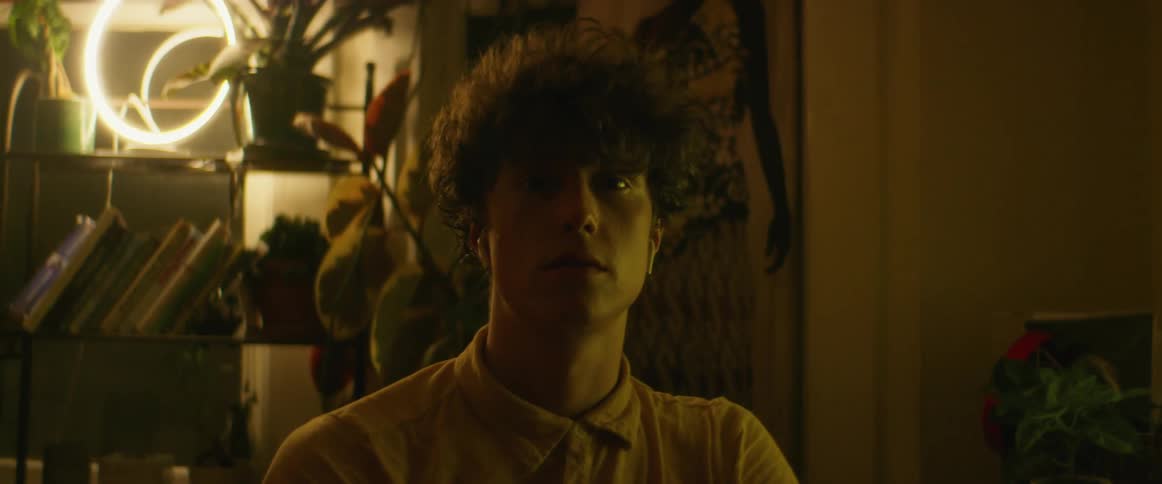 Man with yellow shirt and curly hair sitting at desk uses computer and looks at camera