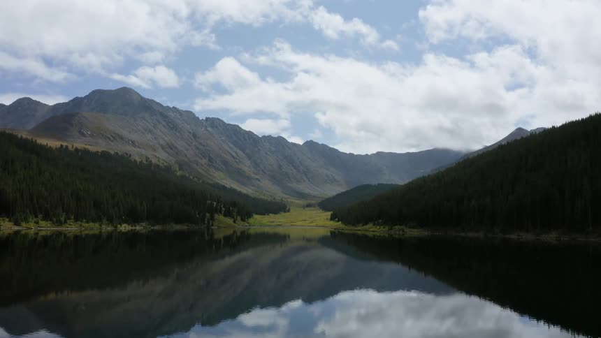An aerial view of approaching the grassy shore of a lake surrounded by a forest with mountains in the background