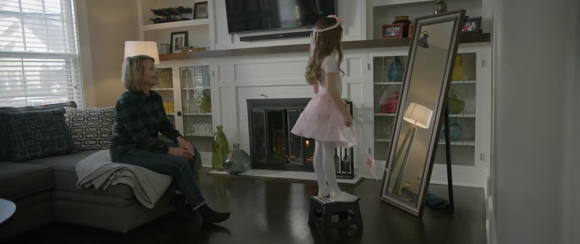 A woman sits as a girl poses with a costume in front of a mirror