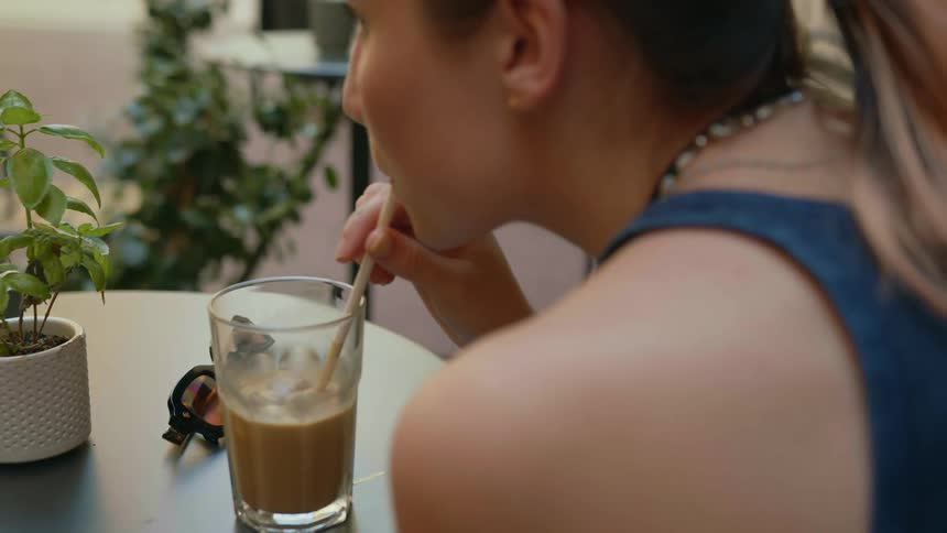 A woman is sitting at a table at an outdoor cafe and sipping a drink through a straw