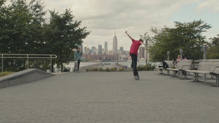 A teenager is riding a skateboard through a city park and does a stunt as someone walks into the park