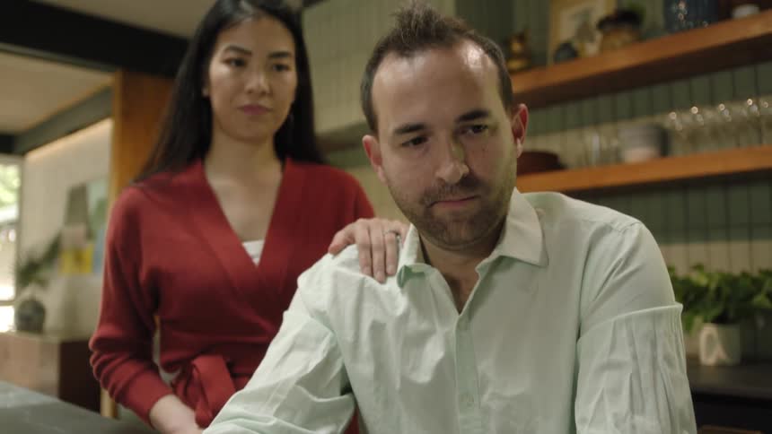 A woman stands behind a man as he moves a bowl of food