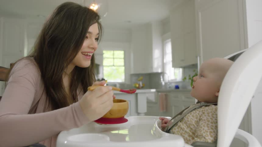 A woman in a residential kitchen is feeding a baby with a spoon in a high chair in the daytime