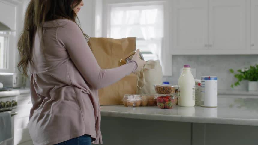 A woman in a kitchen taking groceries out of a brown paper bag with a window and a wall in the background