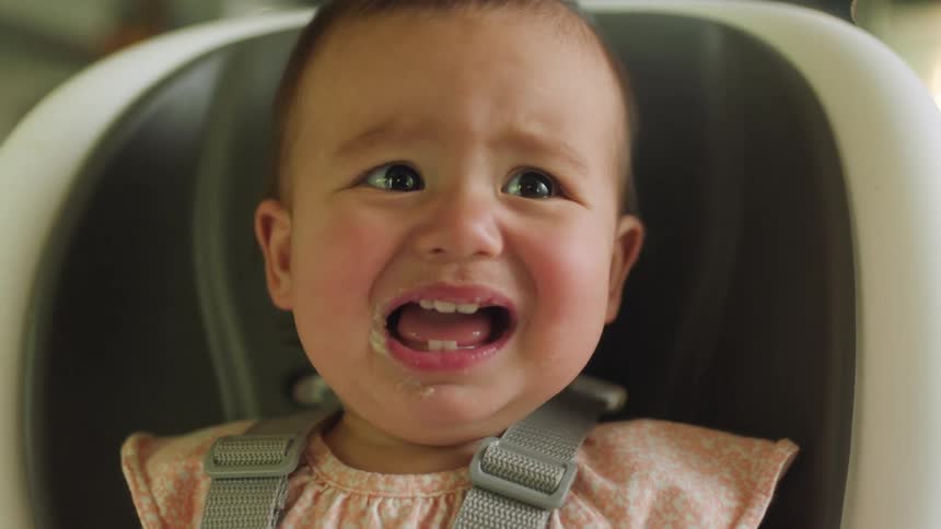 A baby sitting in a high chair while in a home begins crying after reaching for something