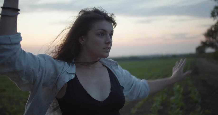 A woman is dancing outdoors near crops on farmland on a breezy evening