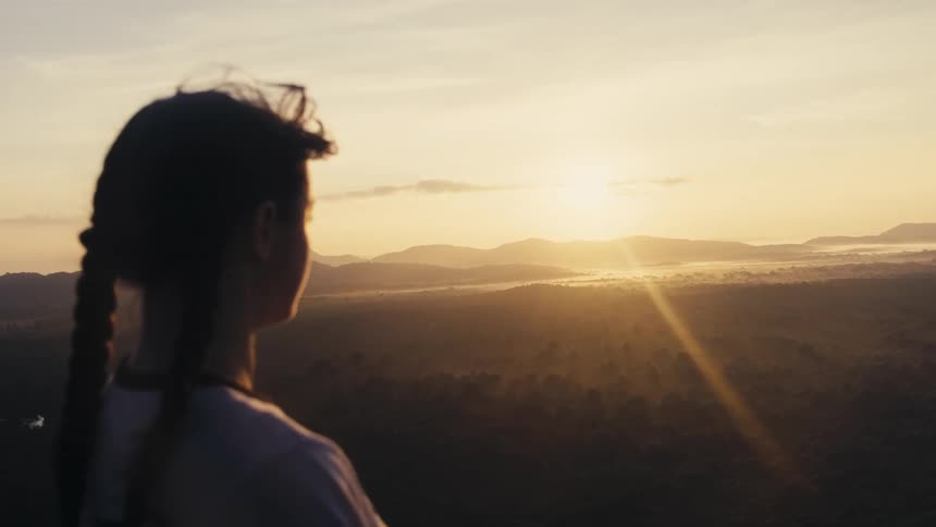 Young woman stands looking across landscape as sun shines from horizon across foggy terrain