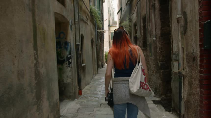 A woman carrying a camera is walking on a stone path in and alley between old buildings