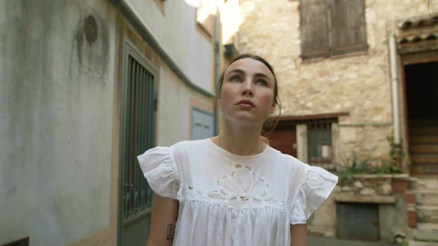 A woman wearing a white dress is walking in an alley looking at buildings
