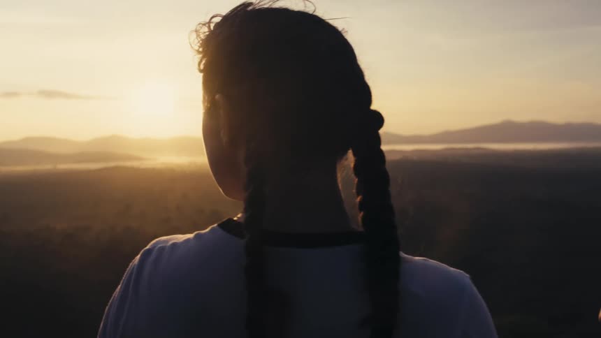 Woman with braids standing on edge of cliff watches the horizon and sunset