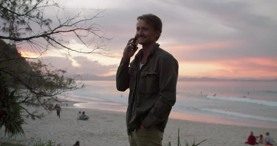 A man talking on a cell phone while standing on a beach at sunset