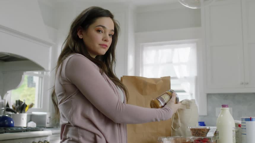 A woman is taking groceries out of a brown bag sitting on a kitchen counter