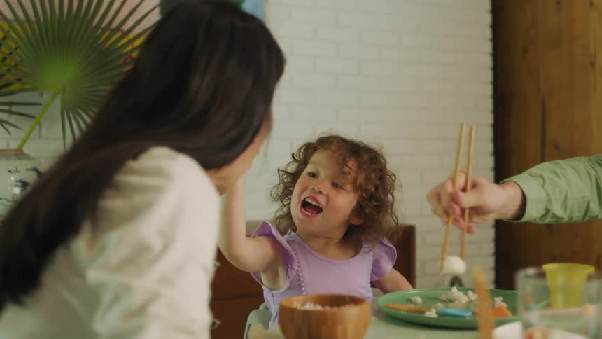A father puts some food on a little girls plate and she picks it up with chopsticks and feeds it to her mother
