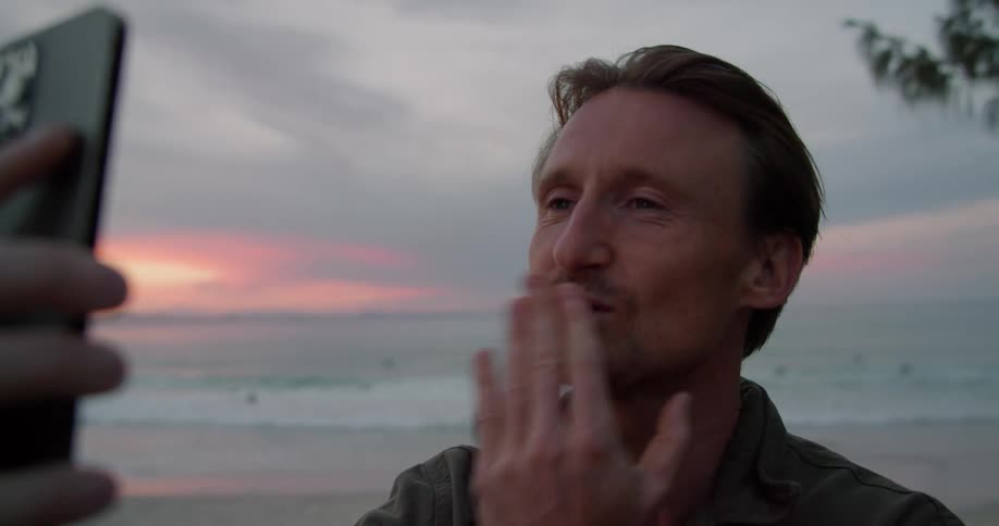 A man is standing on a beach holding his cell phone and having a video conversation as he blows them a kiss