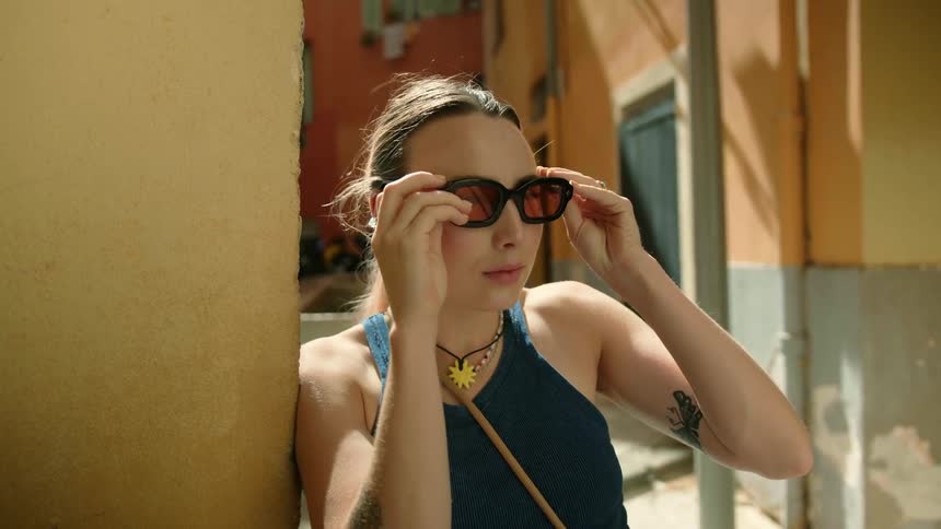 A woman leans against the wall of a building as she takes her sunglasses off of her shirt and puts them on and looks around