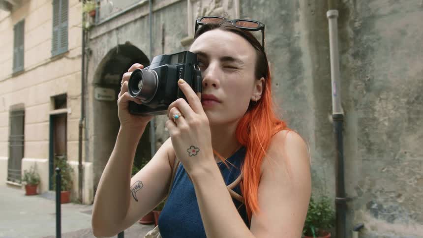 Woman with bright red hair takes picture as pedestrian walks along sidewalk in background