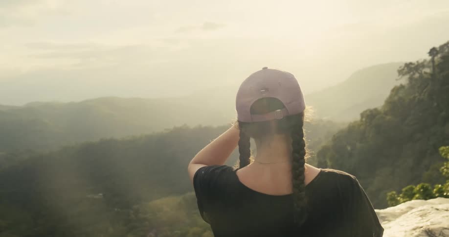 Woman wearing purple baseball cap stands looking over mist covered forested valley surrounded by mountains