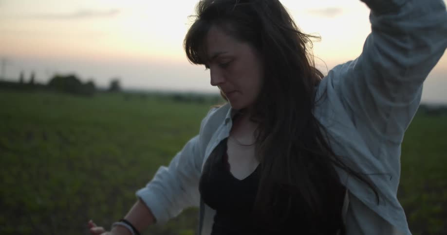 A woman is dancing and swaying in a green field in the country at dusk