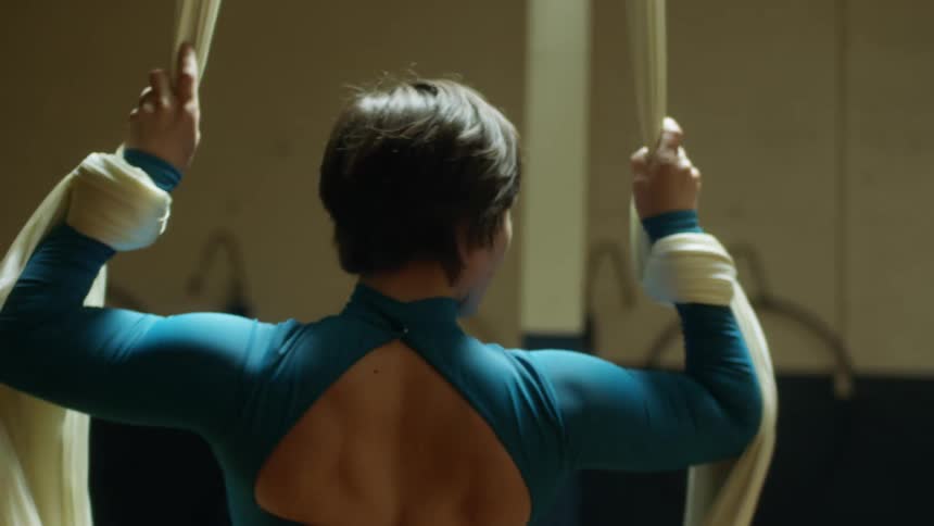 A woman in a gym sitting on a swing made of cloth with a wall and pillar in the background