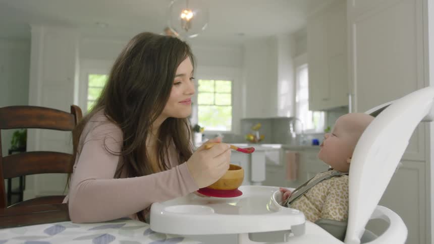 A woman at home smiles as she spoon feeds her baby