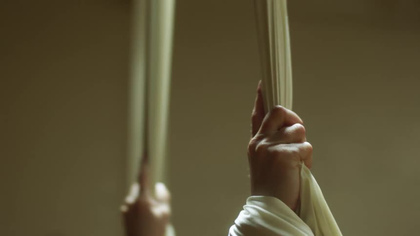 A close view of the hands of a dancer gripping onto fabric as she dangles above the floor slowly spinning while in a dance studio at night