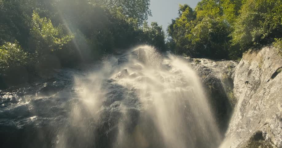 Water flows down rough uneven incline in forest in series of falls spraying water into air and creating rainbow