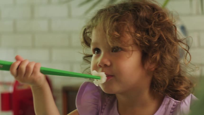 A person helping a little girl in a room eat rice with chopsticks with a plant and a wall in the background