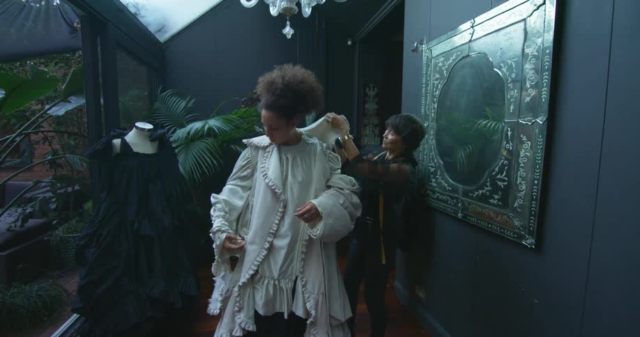 A woman with a measuring tape fixing a dress on another woman with a wall a doorway and a plant in the background