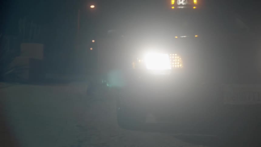 A vehicle approaches and parks on the side of the street at night with bright headlights glaring