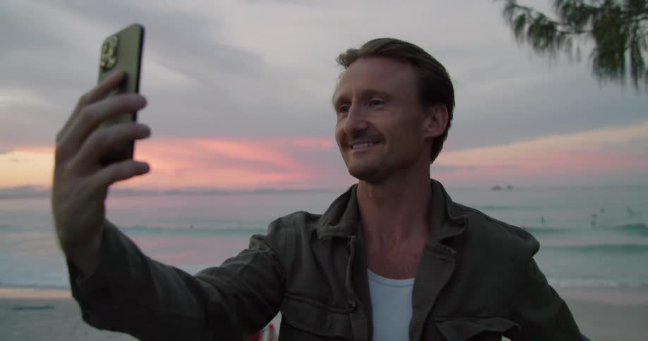 A man holds his phone up for a video call while standing on the beach