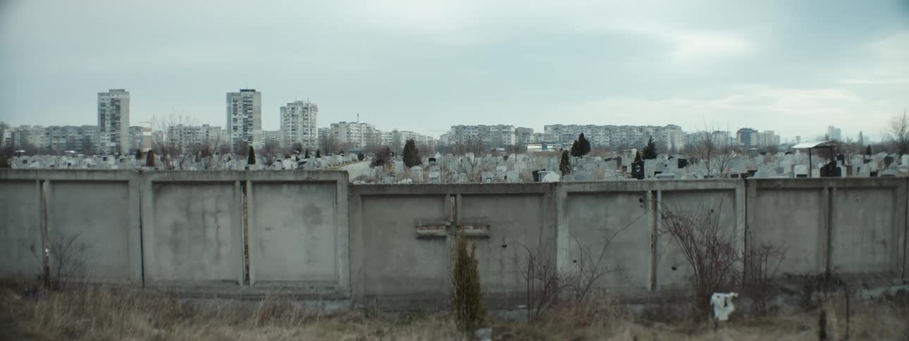 View of graveyard surrounded by wall with cross and city skyline under cloudy sky
