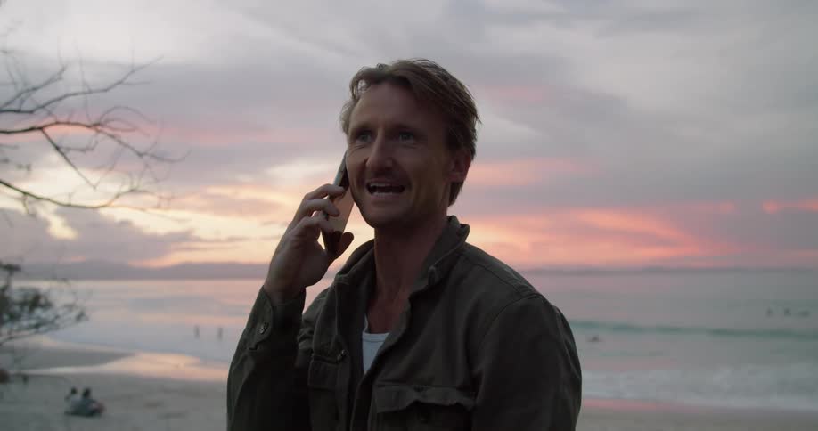 A man laughs while talking on the phone at the beach