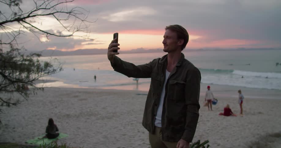 A man is standing video chatting on a smartphone at a populated beach at dusk