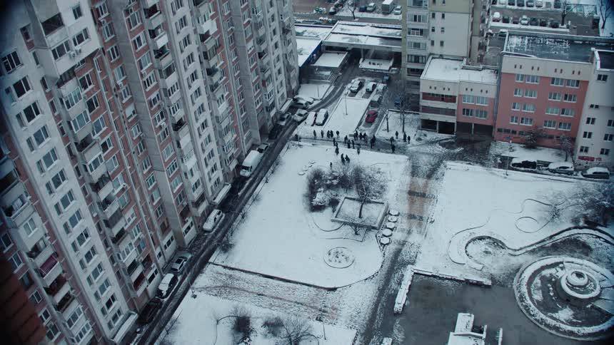 Aerial view of people standing near a snow covered park across from tall apartment buildings and a car traveling on the street