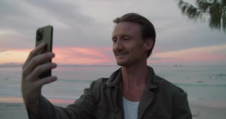 A man is standing on a beach holding his cell phone and having a video conversation