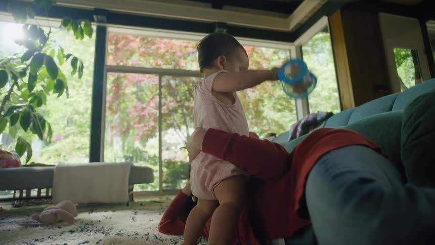 Mother helps baby stand while baby plays with toy as mother lies on floor in living room