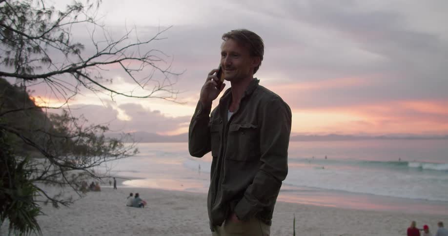 Blonde man with long sleeve drab shirt talks on smartphone next to the beach at sunset