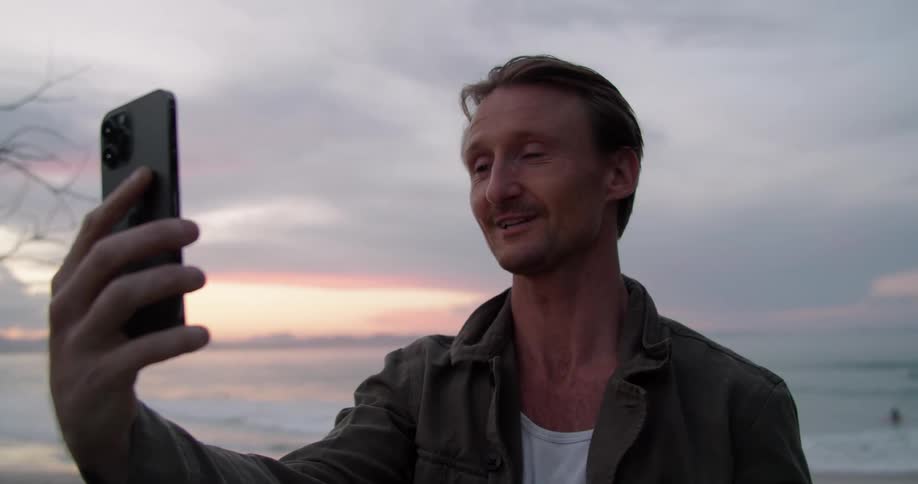 A man stands on a beach looking into a cellphone and blows a kiss