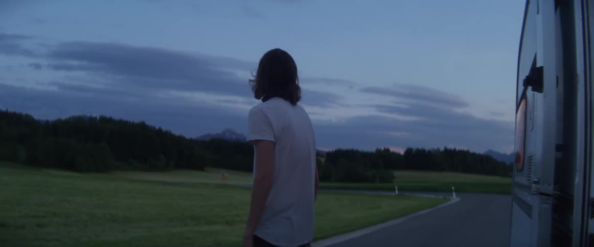 The door to a trailer opens and a young man wearing a white shirt gets out and looks around at the nearby forest