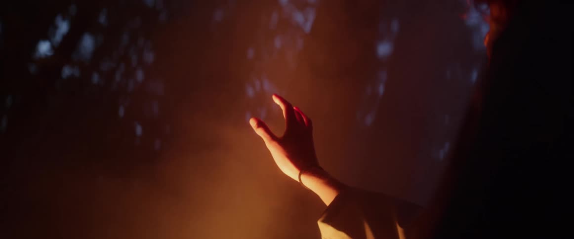 Man with long hair and jacket reaches his hand out to smoky campfire light in the dark forest