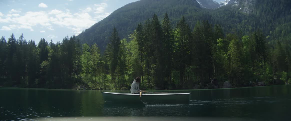 A person paddling a rowboat on a lake with trees a mountain range the sky and clouds in the background