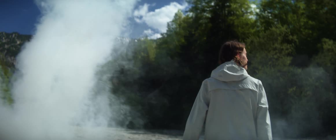 A cloud of steam billows past a man walking and looking around at the trees on a bright blue sky day