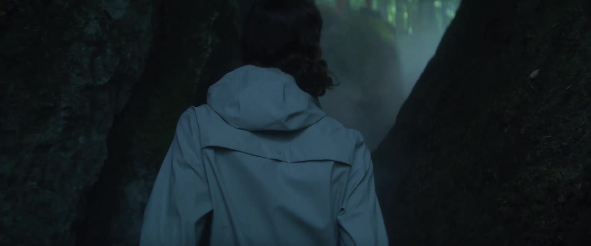 A woman is making her way through a narrow passageway below ground level in a forest with fog during the day
