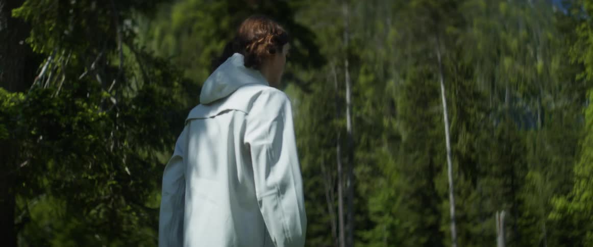A young man wearing a light colored hooded jacket walks into a forest and looks around