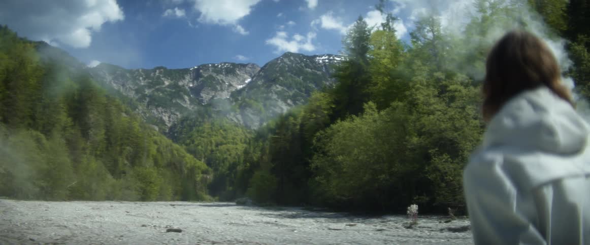 A man walking a landscape with smoke with trees a mountain range the sky and clouds in the background