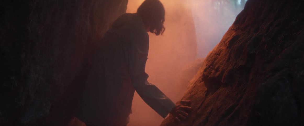 Man with long hair and grey coat walks between high rocks to the light in the foggy forest at dusk