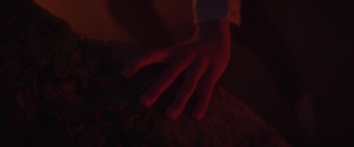 Someone inside a cave is putting their hand on rocks in the darkness and a light shines outside the cave