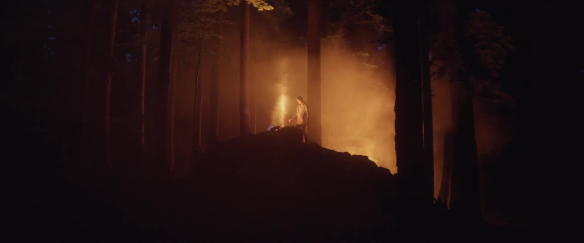A man climbs a structure in the woods at night that has smoke rising from a chimney