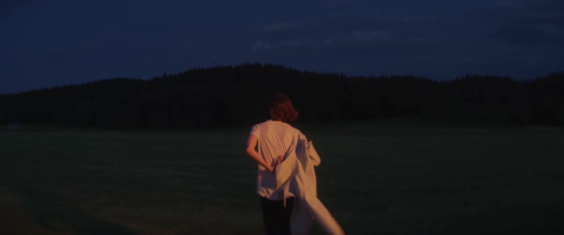 A man puts on a hoodie as he begins to walk across a field towards a mountain at night